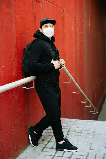 un joven en ropa deportiva negra y una gorra de béisbol con una máscara médica en la cara cerca de la pared roja. protección contra el virus
 - Foto, Imagen
