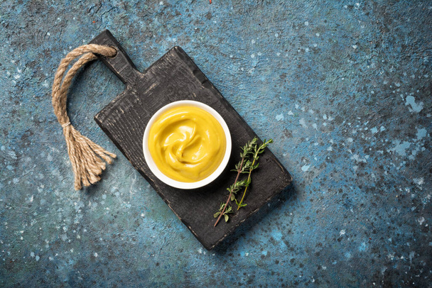 Top view of yellow homemade mustard sauce in small white bowl on black wooden board and blue concrete background - Zdjęcie, obraz