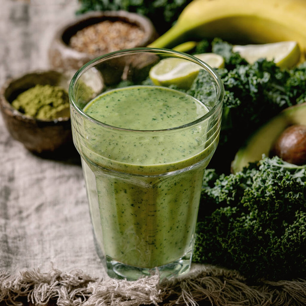 Glass of green healthy vegan smoothie, glass straw. Ingredients above. Kale, bananas, avocado, lime, non-diary milk, matcha powder and seeds over wooden plank cloth background. Close up - Zdjęcie, obraz