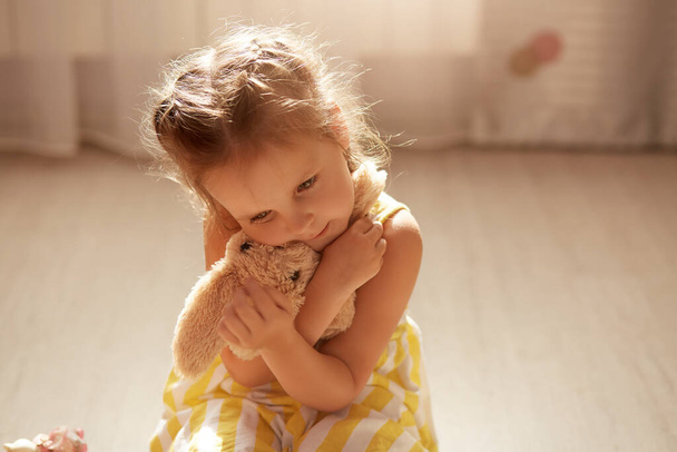 Cute upset little girl wearing yellow and white dress sits on floor with her soft toy in hands and hugs it, female child sitting on floor. Self isolation during coronavirus pandemic. Covid 19 concept. - Фото, изображение