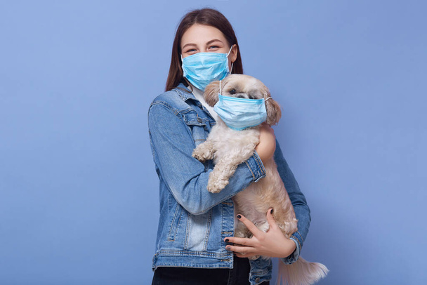 Indoor shot of woman dresses denim jacket protective flumask holds dog pet in medical mask. Coronavirus disease dangerous for people and pets. Coronavirus, pandemic, qaurantine, precautions concept. - Fotoğraf, Görsel