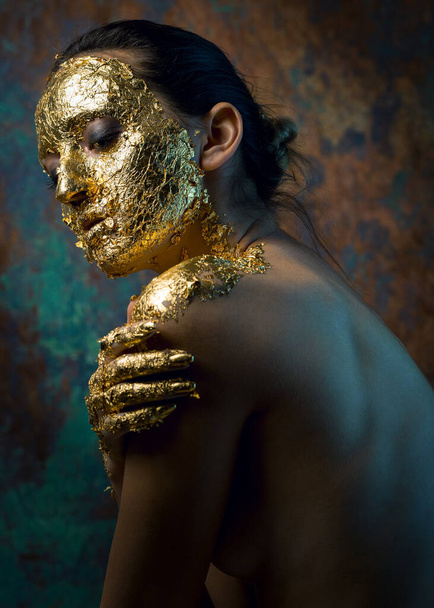 Girl with a mask on her face made of gold leaf. Gloomy studio portrait of a brunette on an abstract background. - Fotó, kép