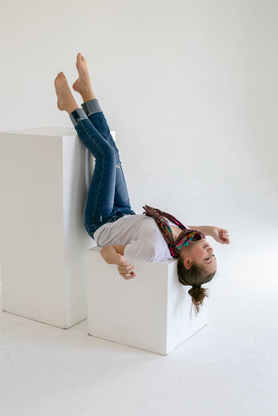Босоногая девушка-подросток в синих джинсах лежит вверх ногами на белом кубе в белой студии
 - Фото, изображение