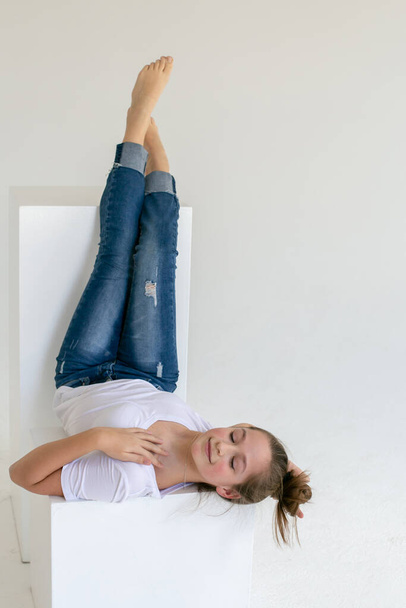slender barefoot girl in blue jeans and a white t-shirt lies upside down on a white cube in a white studio - Фото, изображение