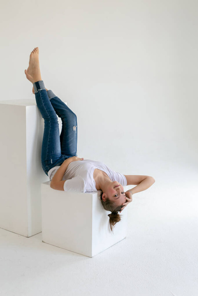 barefoot teenager girl in blue jeans and a white t-shirt lies upside down on a white cube in a white studio - Фото, зображення