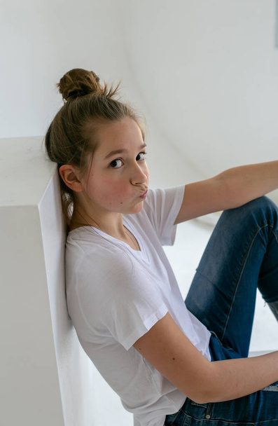 zamyšlená mladá dívka v modrých džínách a bílém tričku sedí na bílé kostce v bílém studiu a dívá se do kamery - Fotografie, Obrázek