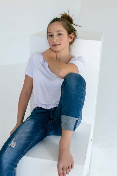 Mavi kotlu ve beyaz tişörtlü tatlı genç kız beyaz bir küpün üzerinde oturuyor ve kameraya bakıyor. - Fotoğraf, Görsel