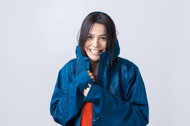 Portrait of a smiling girl dressed in blue raincoat in drops posing with hood on grey background in a studio. - Photo, Image