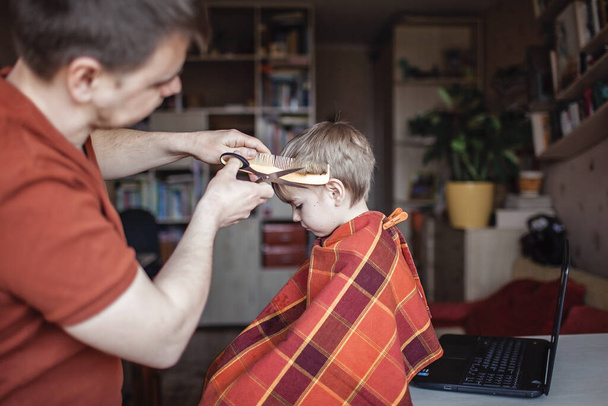 中世の父カッティング毛に彼の小さな息子に自分自身によってホーム見ますビデオ放送でラップトップ,髪型オンラインクラス,滞在家と生活中にロックダウン - 写真・画像
