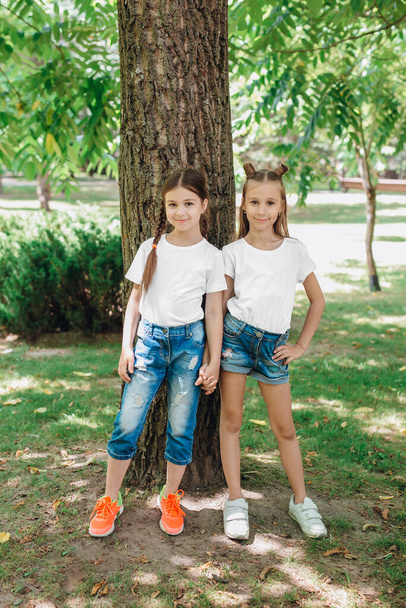 Two little girls in white t-shirts standing in park outdoor. Mock up. - Φωτογραφία, εικόνα