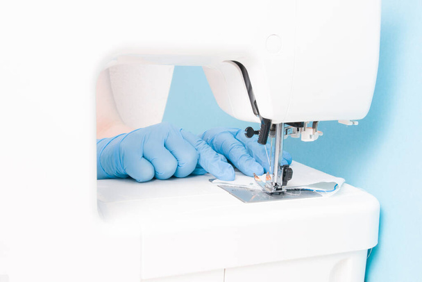 a woman in blue disposable medical gloves sews on a sewing machine a protective face mask made of cotton gray fabric, blue background, the process of sewing masks - Fotoğraf, Görsel