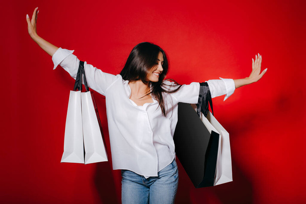 Mujer feliz mostrando sus nuevas compras, sosteniendo grandes bolsas de compras en dos manos sobre fondo rojo
. - Foto, imagen