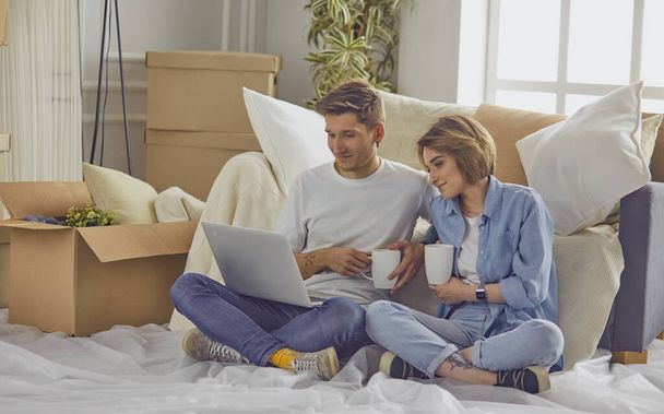 Portret van een gelukkig stel op zoek naar laptop computer samen zitten in nieuw huis, omgeven met dozen - Foto, afbeelding
