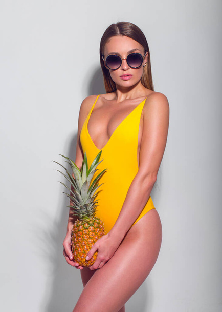 Sexy braun gebranntes schlankes Mädchen in gelbem Badeanzug und Sonnenbrille posiert mit Ananas in den Händen auf weißem Hintergrund - Foto, Bild
