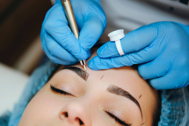 Permanent make-up for eyebrows of beautiful brunette woman in beauty salon. Closeup beautician in gloves doing microblading for eyebrow. - Фото, зображення