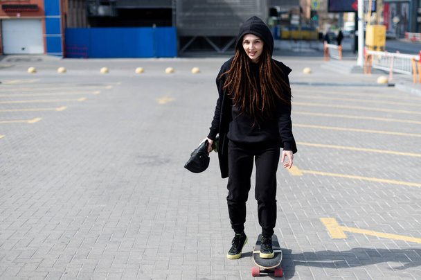 Cute girl with dreadlocks in a hood rides on a skateboard. girl in black clothes with a gas mask in hand - Photo, image