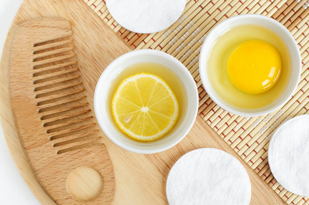 Lemon juice with lemon slice, raw egg in the small white bowls and wooden hair brush. Natural homemade hair treatment and zero waste concept. Top view, copy space - 写真・画像