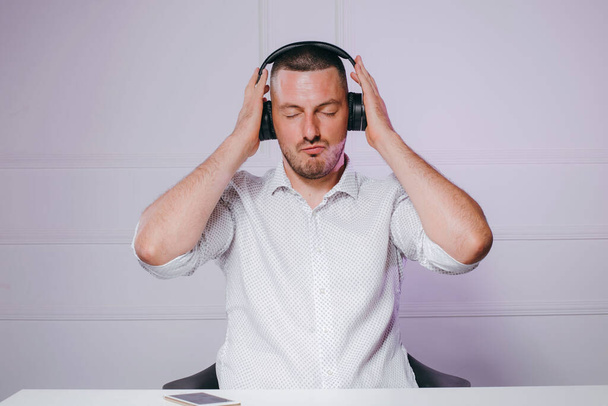 Young man with closed eyes in white shirt and wireless headphones sitting at the table, listening to music. - Photo, Image
