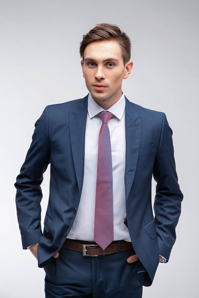 Hombre joven y guapo con el pelo corto oscuro en un traje azul en una camisa blanca con una corbata sobre un fondo blanco en el estudio
 - Foto, imagen