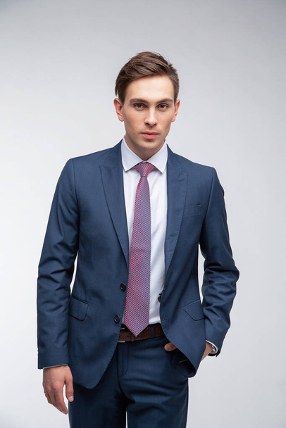 Młody, przystojny mężczyzna o ciemnych krótkich włosach w niebieskim garniturze w białej koszuli z krawatem na białym tle w studio - Zdjęcie, obraz