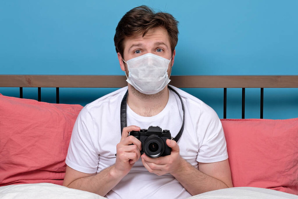 Młody fotograf w masce medycznej chory siedzi sam na kwarantannie z kamerą, odizolowany na niebieskiej ścianie. - Zdjęcie, obraz
