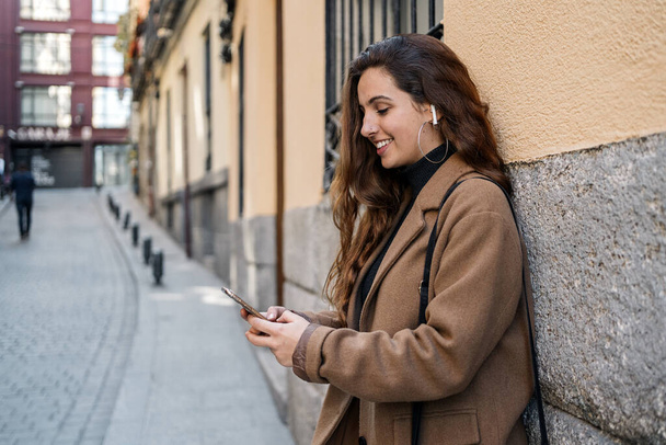 Archivbild einer jungen Kaukasierin mit einem Smartphone auf der Straße. Sie lächelt. Sie trägt schicke Freizeitkleidung. - Foto, Bild