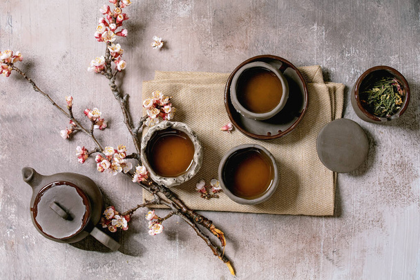 Tea drinking wabi sabi japanese style dark clay cups and teapot on cloth napkin with blooming cherry branches. Grey texture concrete background. Flat lay, space - Photo, Image