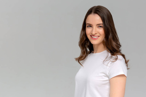 Nahaufnahme Porträt einer lächelnden jungen Frau in weißem T-Shirt, die in die Kamera blickt, isoliert auf grauem Hintergrund - Foto, Bild