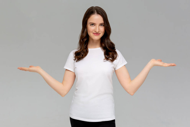 Close up portrait of smiling young woman in white t-shirt throw their hands up looking at camera, isolated on gray background - Photo, image