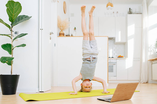 Cute little boy doing head stands, looking at laptop. Physical athletic exercise on isolation at home. - Foto, afbeelding
