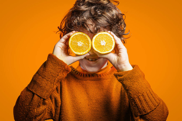 Το αγοράκι που κρατάει ένα πορτοκάλι στα χέρια του, καλύπτει τα μάτια του με πορτοκαλί φόντο. Διατροφή και βιταμίνη C έννοια της υγείας. - Φωτογραφία, εικόνα