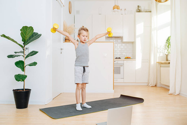 Little boy exercising with dumbbells, holding them in arms extended to side. Improving strength and average fitness while staying on quarantine. - Fotoğraf, Görsel