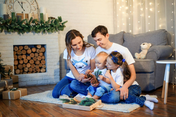 Theme Christmas and New Year family circle. Young Caucasian family with 1 year old son and 6 years old daughter sitting on floor in living room. Christmas decorations. Boy and girl kids and parents. - Foto, Bild