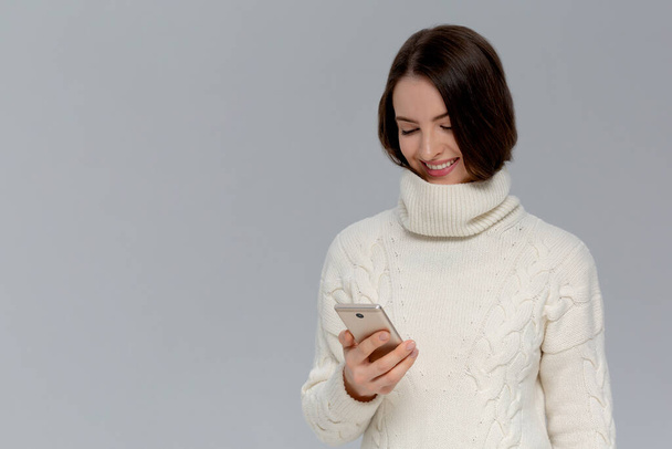 Joyful emotive young european woman in a white sweater. holding smartphone and smiling cheerfully at camera while retelling interesting article to friend - Photo, Image