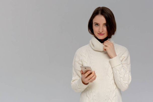 Gioiosa giovane donna europea emotiva con un maglione bianco. tenendo smartphone e sorridendo allegramente alla fotocamera mentre raccontava un articolo interessante ad un amico
 - Foto, immagini