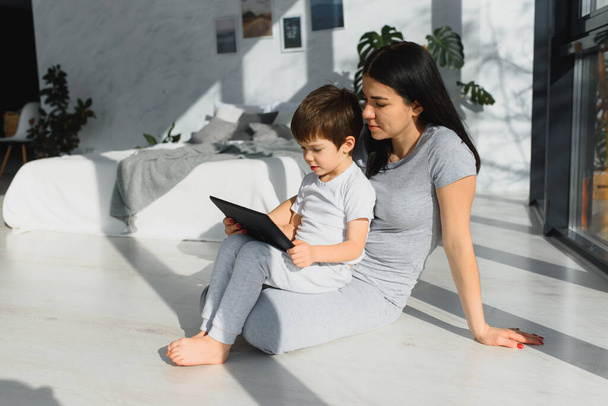 Cute little boy and his young mother using a digital tablet and smiling on the floor at home. Happy family with gadget - Φωτογραφία, εικόνα