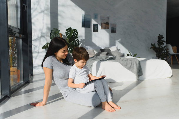 Sevimli küçük çocuk ve genç annesi dijital tablet kullanıyorlar ve evde yerde sırıtıyorlar. Aletli mutlu aile - Fotoğraf, Görsel