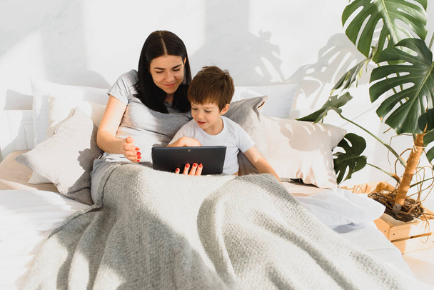 Piękna uśmiechnięta matka i syn bawią się cyfrowym tabletem leżącym na łóżku. - Zdjęcie, obraz