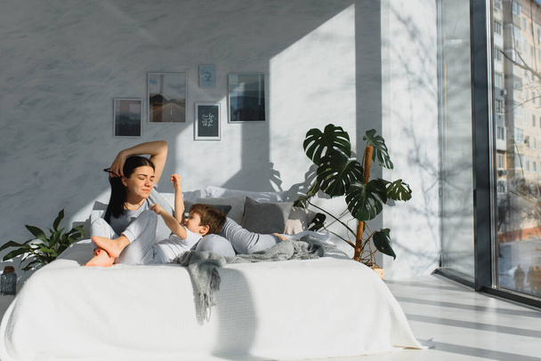 Young mother with her 4 years old little son dressed in pajamas are relaxing and playing in the bed at the weekend together, lazy morning, warm and cozy scene. - Фото, изображение
