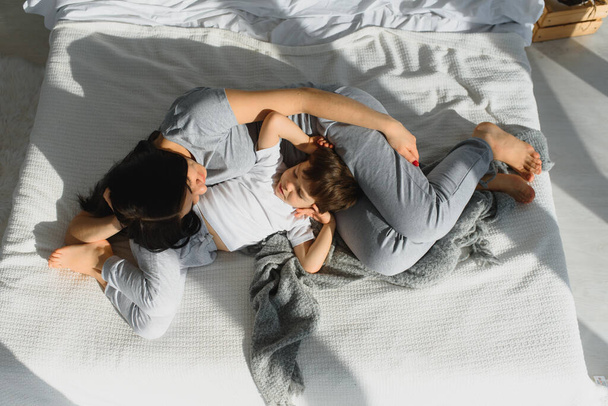 Young mother with her 4 years old little son dressed in pajamas are relaxing and playing in the bed at the weekend together, lazy morning, warm and cozy scene. - Foto, imagen