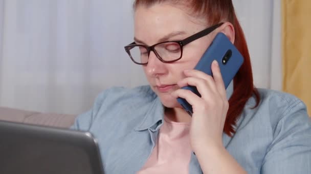Портрет втомленої молодої жінки, що працює на ноутбуці, що сидить у вітальні. Жінка розмовляє на мобільному телефоні
. - Кадри, відео
