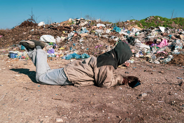 Βρώμικη άστεγη βρίσκεται στο έδαφος κοντά σε χωματερή και κοιτάζει μέσα στην ταμπλέτα. - Φωτογραφία, εικόνα