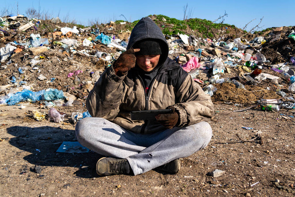 汚れたホームレスの女性が埋め立て地の近くの地面に座ってタブレットを見て - 写真・画像