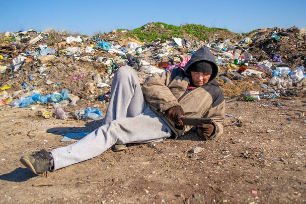 Dirty homeless woman lies on the ground near landfill and looks into the tablet - Fotografie, Obrázek
