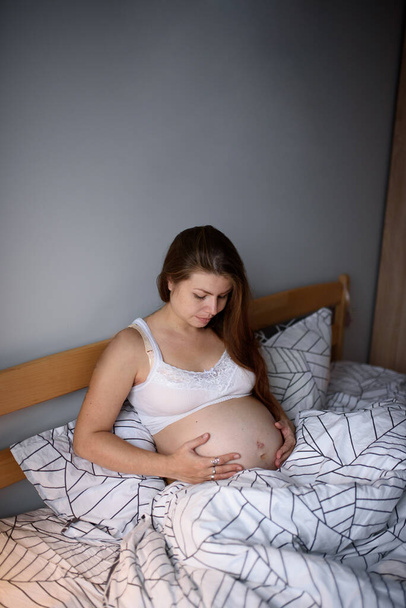 A pregnant girl clings to her belly and worries before giving birth during a krantin. Self-isolation at home. - Foto, imagen
