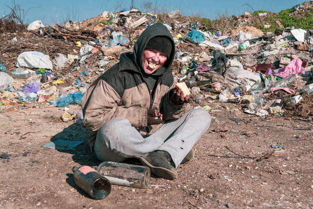 Una sporca senzatetto si siede a terra vicino a una discarica e mangia
 - Foto, immagini