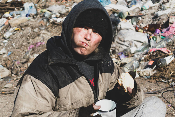 Eine schmutzige Obdachlose sitzt auf dem Boden in der Nähe einer Mülldeponie und isst - Foto, Bild