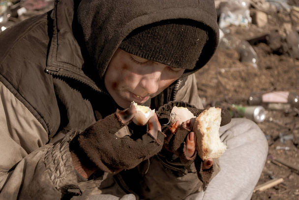 Una sucia indigente se sienta en el suelo cerca de un vertedero y come
 - Foto, Imagen
