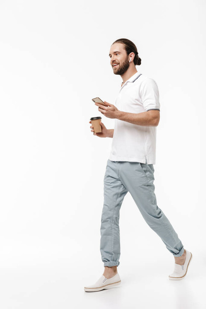 Photo of a young positive happy handsome bearded man walking isolated over white wall background listening music with earphones drinking coffee using mobile phone. - Fotografie, Obrázek