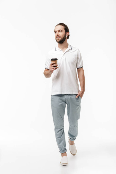 Photo of a young positive happy handsome bearded man walking isolated over white wall background listening music with earphones drinking coffee. - Foto, Imagem